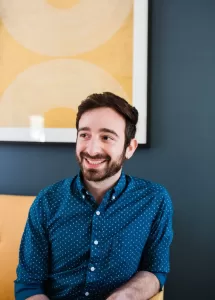 Frank Molignano, a homeopath at The Healing Narrative, seated on a yellow couch against a dark blue wall with a yellow picture behind him, looking to the right